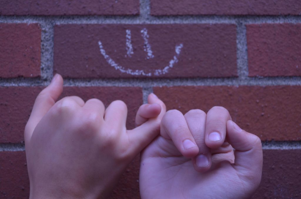 Two hands making a pinkie-promise in front of a chalk smiley face drawn on a brick wall.